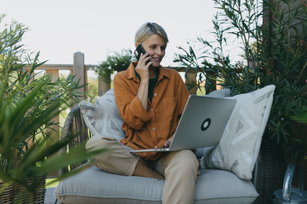 Woman working in garden, with laptop on legs. Businesswoman working remotely from outdoor homeoffice, making call on smartphone. Concept of outdoor home office.