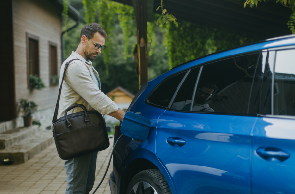 Portrait of businessman charging his electric car in front of his house. Pluging charger in charging port.