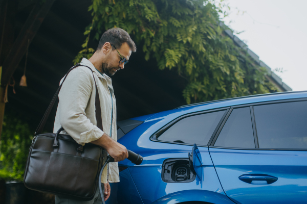 Portrait of a man charging his electric car in front of his house, plugging the charger into the charging port. The man is unplugging the charger from the fully charged car before going to the office.