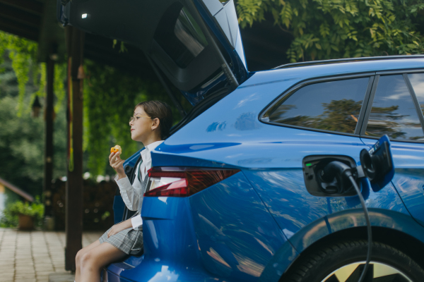 Girl and electric car with charger in charging port. Girl sitting in car trunk, eating apple while vehicle is charging before going to school.