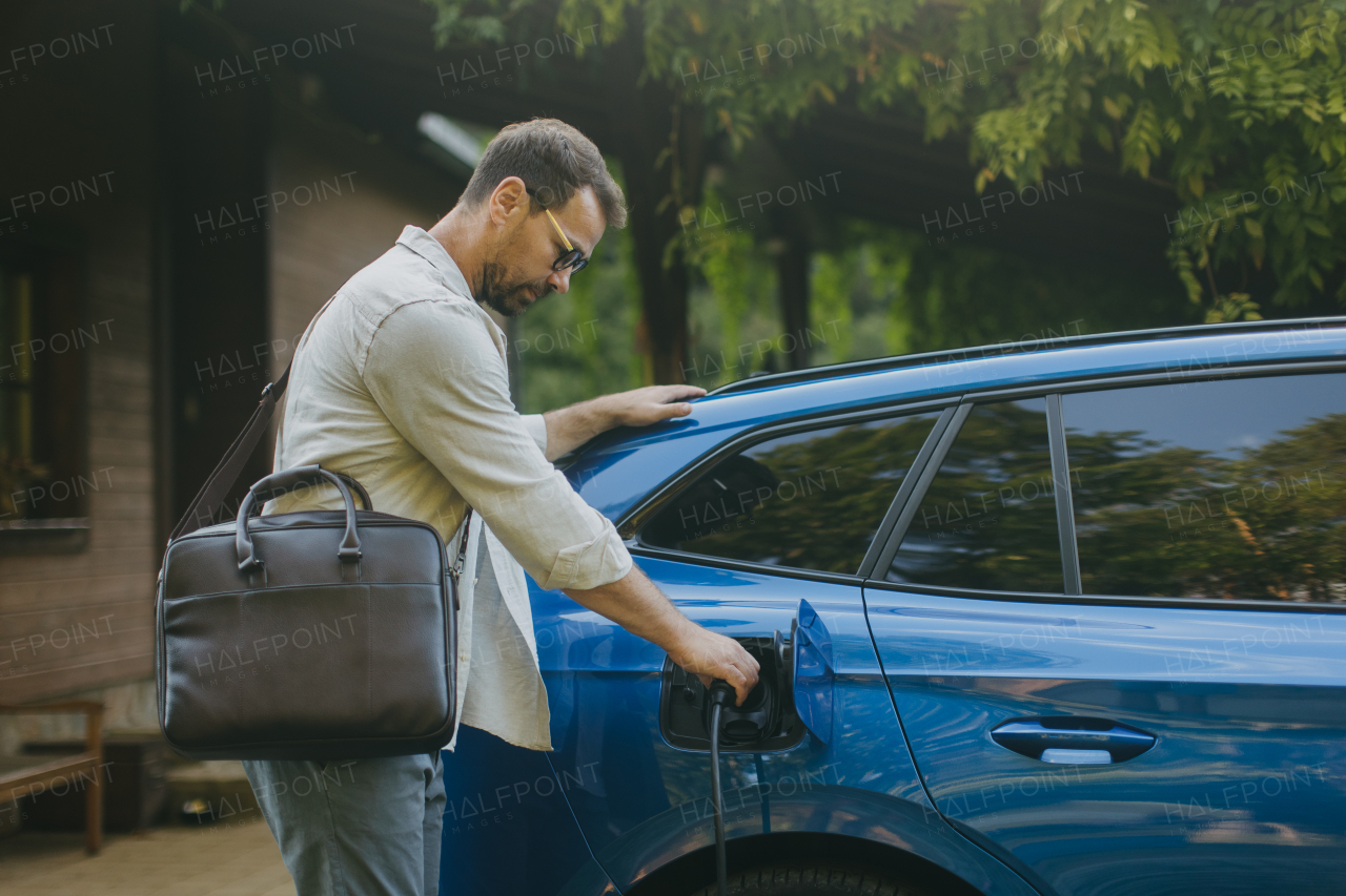 Portrait of a man charging his electric car in front of his house, plugging the charger into the charging port. The man is unplugging the charger from the fully charged car before going to the office.