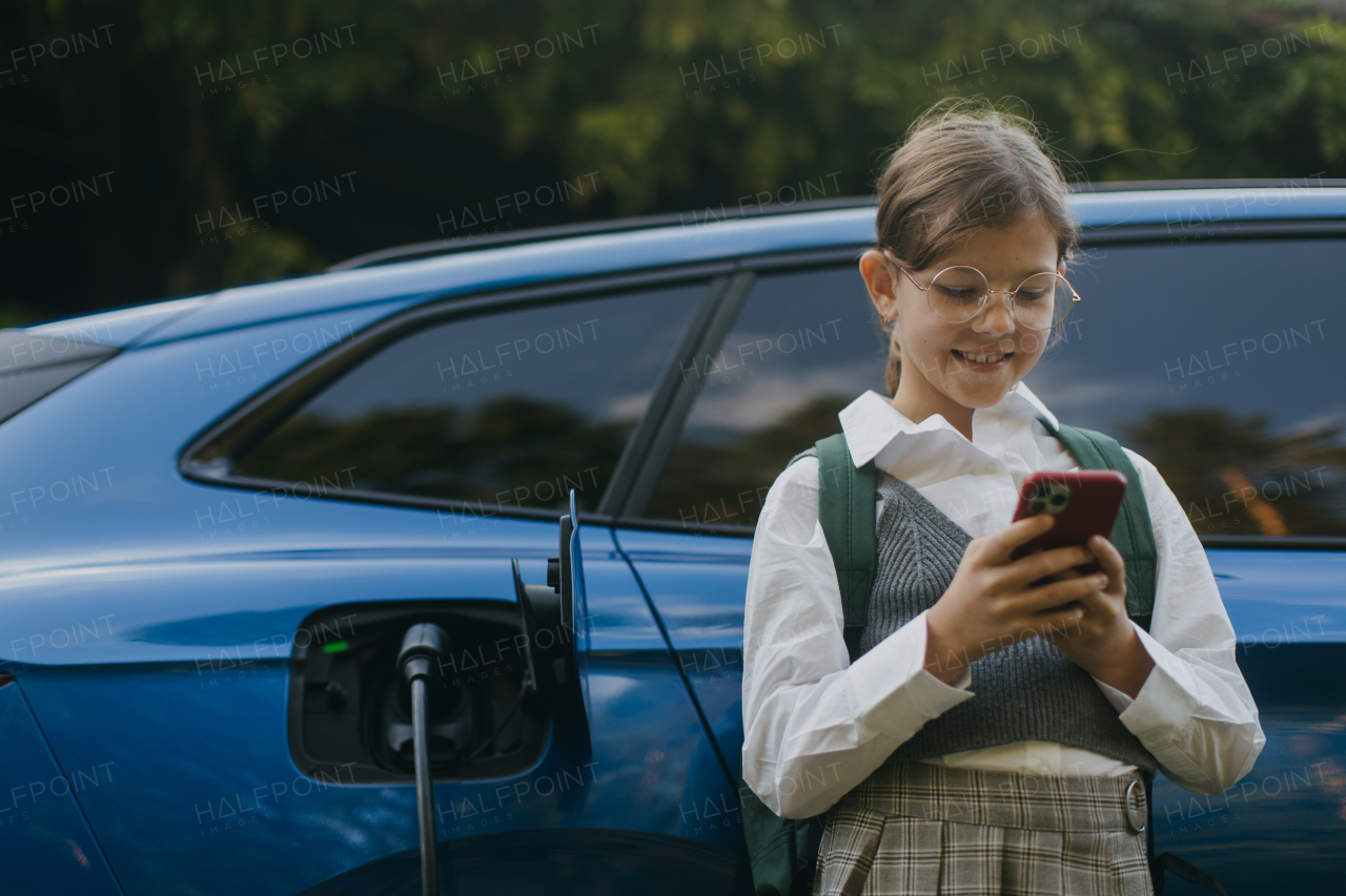 Girl and electric car with charger in charging port. Girl scrolling on her smartphone while care is charging before going to school.