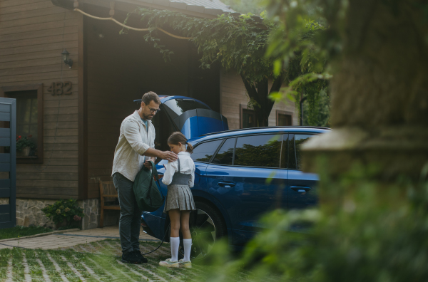 Portrait of a man charging his electric car in front of his house, plugging the charger into the charging port. The man is unplugging the charger from the fully charged car before driving daughter to school.