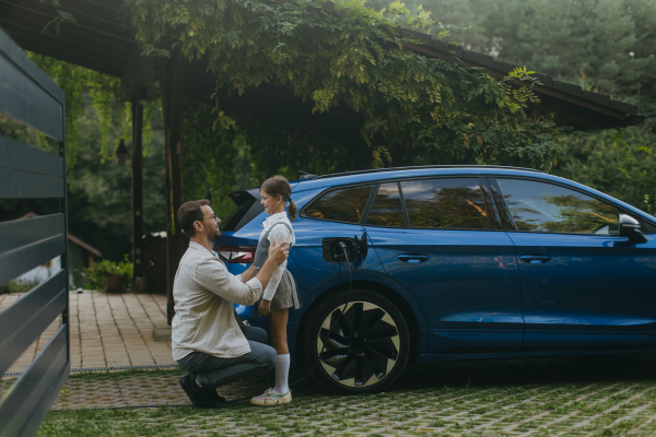 Portrait of father and daughter talking, while charging their family electric car in front of house. Electric vehicle with charger in charging port. Dad driving daughter to school.