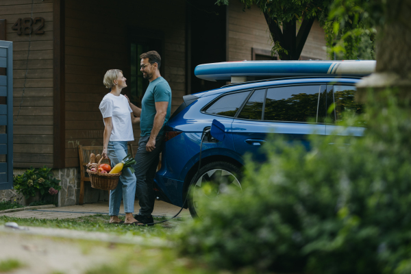 Couple standing by their electric car, looking at each other. Electric vehicle with charger in charging port.