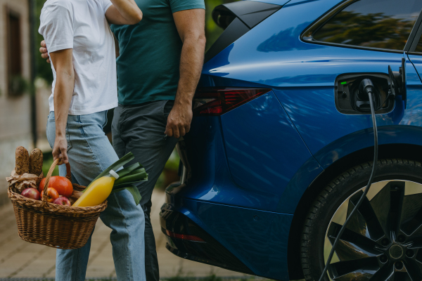 Close-up of couple with groceries in a basket, standing by a charging electric car. Family electric vehicle with charger in charging port.