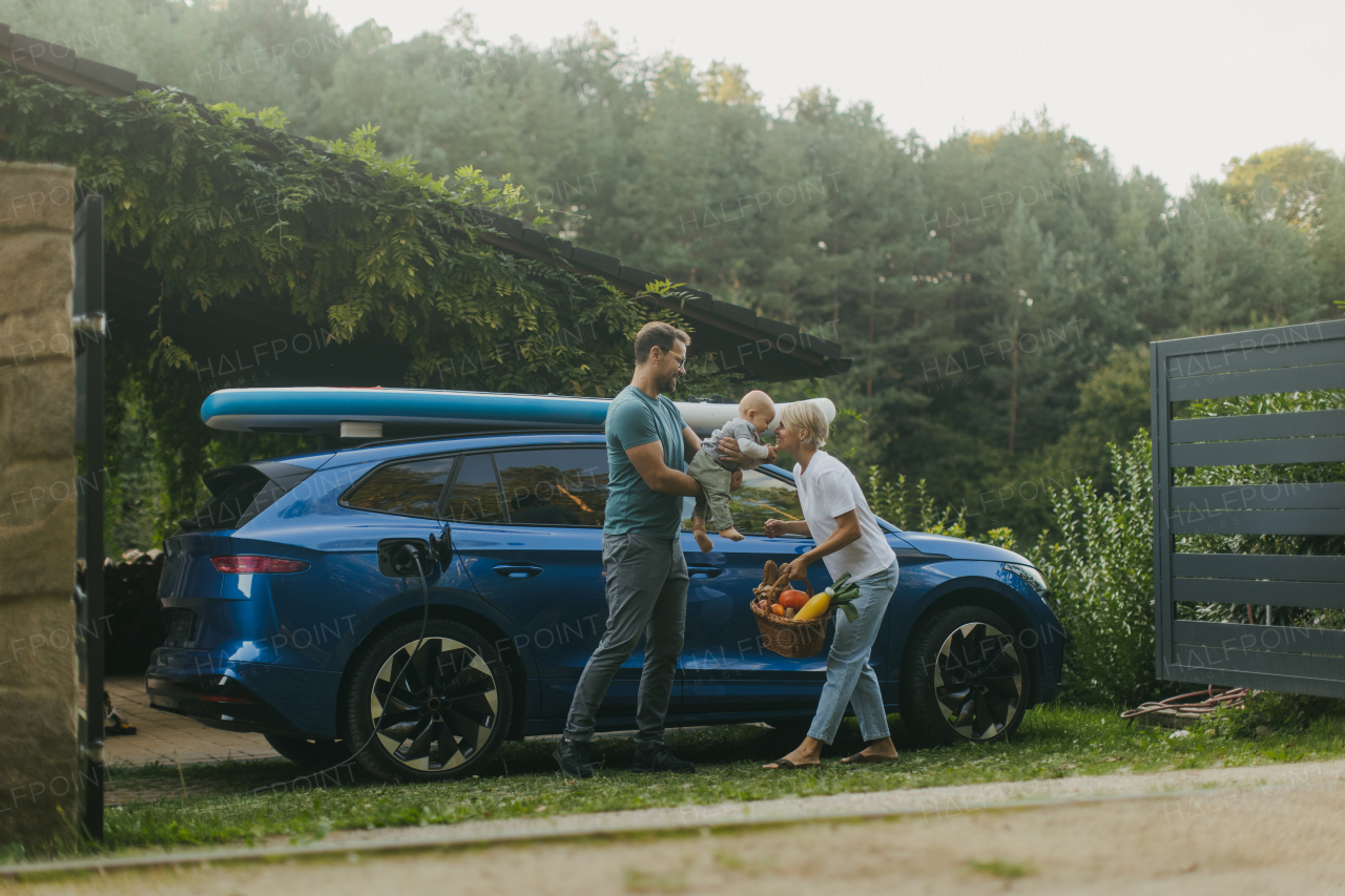 Young family with little baby unloading groceries from their electric car. Electric vehicle with charger in charging port.