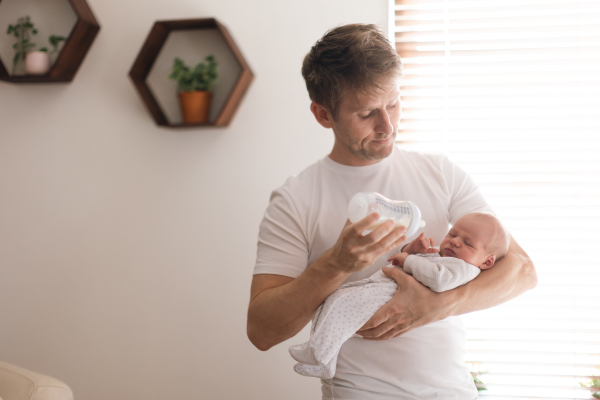 A father holding and feeding his newborn son with milk bottle at home.