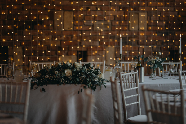 Elegant, romantic table setting for indoor wedding reception with fairy lights on brick wall behind. Luxury wedding table decoration, special event table set up with fresh flowers arragements.