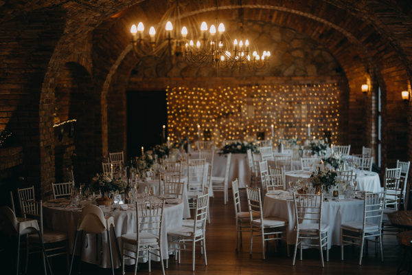 Elegant, romantic table setting for indoor wedding reception with fairy lights on brick wall behind. Luxury winery, brewery cellar wedding venue.