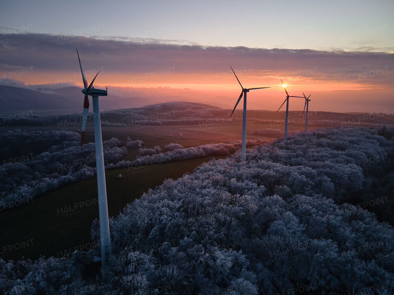 Aerial landscape photography of sunrise over frost-covered nature with wind turbines. Wind turbine towers in soft morning light with icy trees around, harmony of nature and technology. Concept of wind power as clean, renewable energy source.