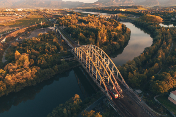 Aerial view of a long train crossing a railway bridge over a river, in autumn nature during day.