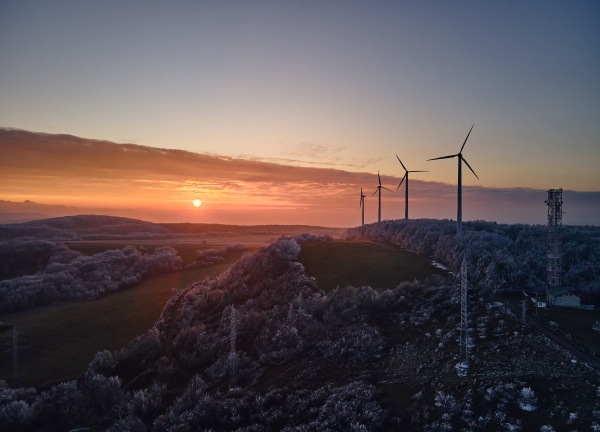 Aerial landscape photography of sunrise over frost-covered nature with wind turbines. Wind turbine towers in soft morning light with icy trees around, harmony of nature and technology. Concept of wind power as clean, renewable energy source.