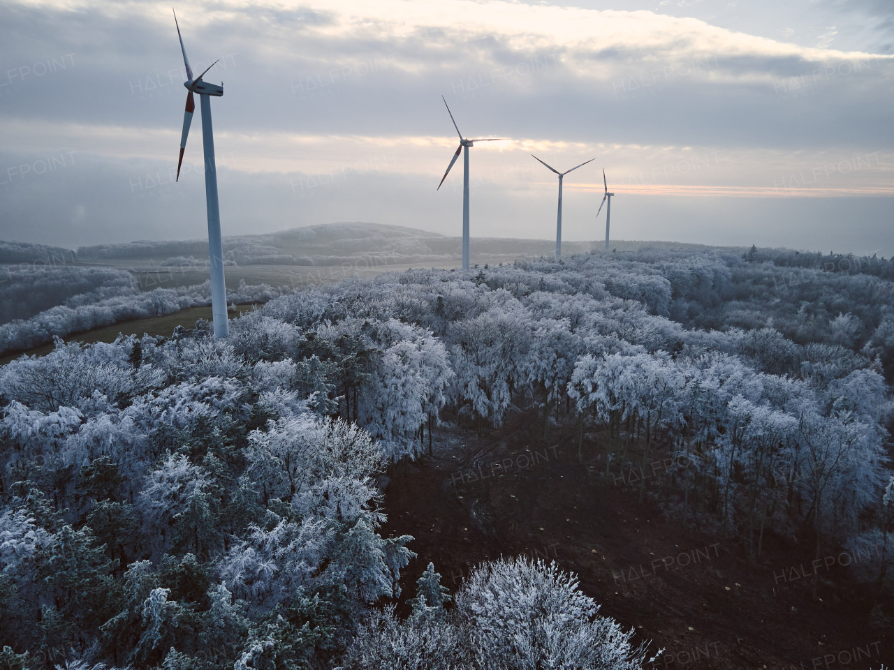 Aerial landscape photography of sunrise over frost-covered nature with wind turbines. Wind turbine towers in soft morning light with icy trees around, harmony of nature and technology. Concept of wind power as clean, renewable energy source.