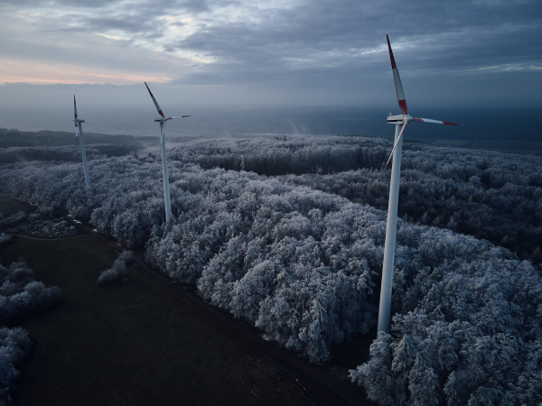 Aerial landscape photography of sunrise over frost-covered nature with wind turbines. Wind turbine towers in soft morning light with icy trees around, harmony of nature and technology. Concept of wind power as clean, renewable energy source.