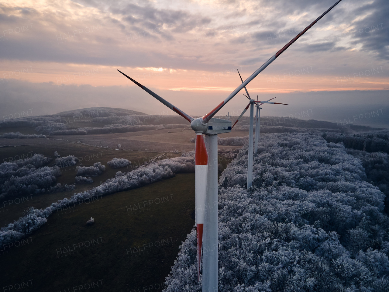 Aerial landscape photography of sunrise over frost-covered nature with wind turbines. Wind turbine towers in soft morning light with icy trees around, harmony of nature and technology. Concept of wind power as clean, renewable energy source.