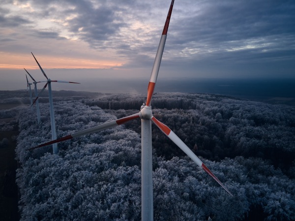 Aerial landscape photography of sunrise over frost-covered nature with wind turbines. Wind turbine towers in soft morning light with icy trees around, harmony of nature and technology. Concept of wind power as clean, renewable energy source.