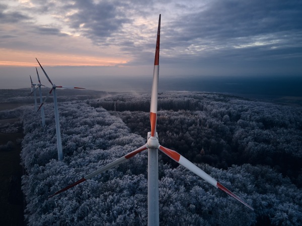 Aerial landscape photography of sunrise over frost-covered nature with wind turbines. Wind turbine towers in soft morning light with icy trees around, harmony of nature and technology. Concept of wind power as clean, renewable energy source.