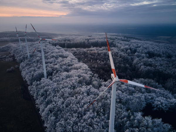 Aerial landscape photography of sunrise over frost-covered nature with wind turbines. Wind turbine towers in soft morning light with icy trees around, harmony of nature and technology. Concept of wind power as clean, renewable energy source.