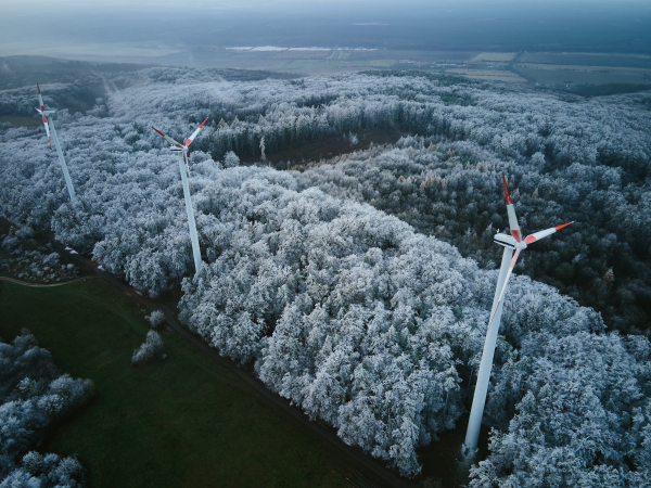 Aerial landscape photography of sunrise over frost-covered nature with wind turbines. Wind turbine towers in soft morning light with icy trees around, harmony of nature and technology. Concept of wind power as clean, renewable energy source.