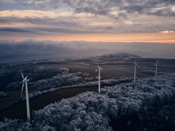 Aerial landscape photography of sunrise over frost-covered nature with wind turbines. Wind turbine towers in soft morning light with icy trees around, harmony of nature and technology. Concept of wind power as clean, renewable energy source.