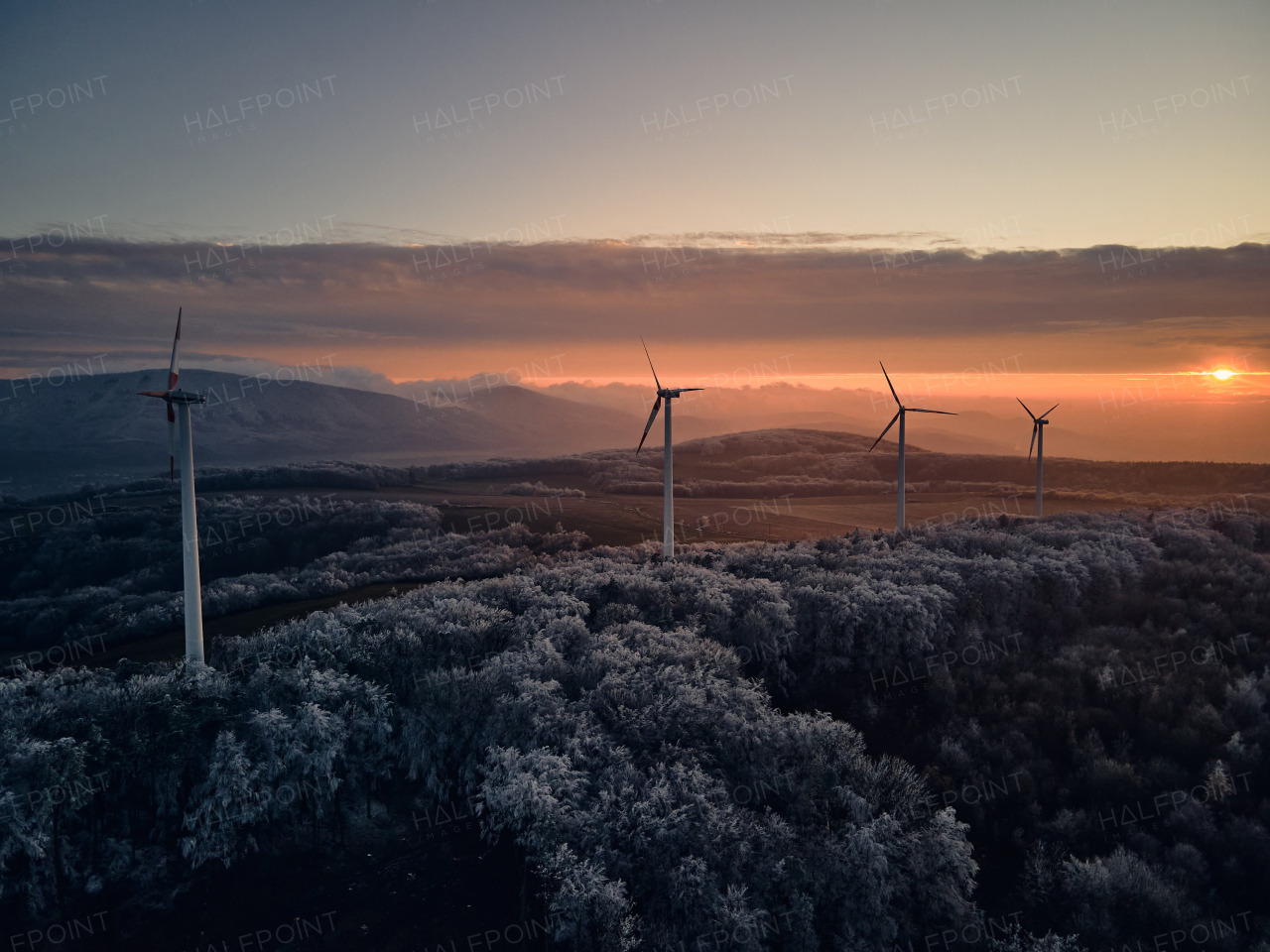 Aerial landscape photography of sunrise over frost-covered nature with wind turbines. Wind turbine towers in soft morning light with icy trees around, harmony of nature and technology. Concept of wind power as clean, renewable energy source.