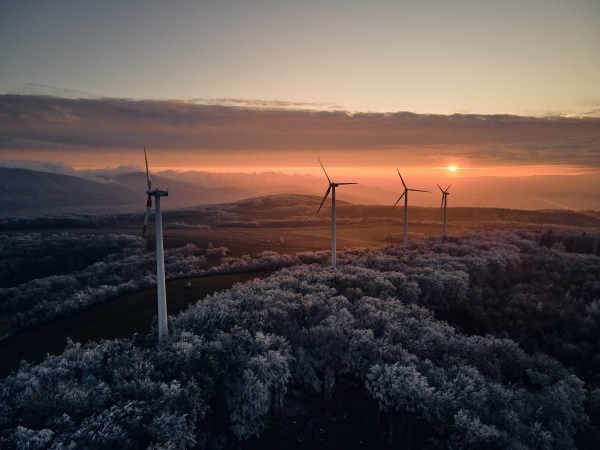 Aerial landscape photography of sunrise over frost-covered nature with wind turbines. Wind turbine towers in soft morning light with icy trees around, harmony of nature and technology. Concept of wind power as clean, renewable energy source.