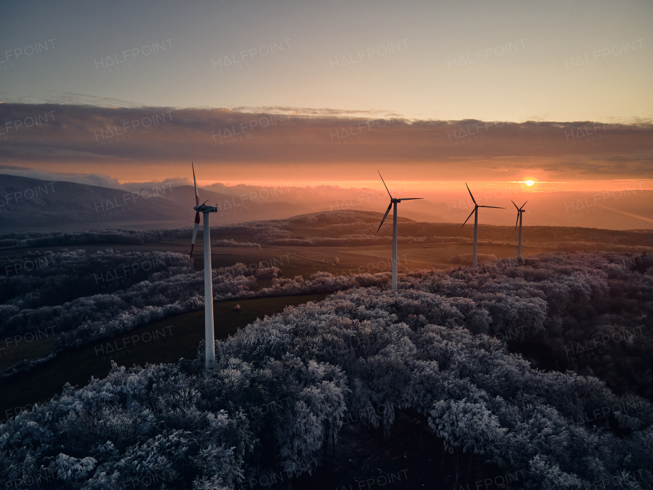 Aerial landscape photography of sunrise over frost-covered nature with wind turbines. Wind turbine towers in soft morning light with icy trees around, harmony of nature and technology. Concept of wind power as clean, renewable energy source.