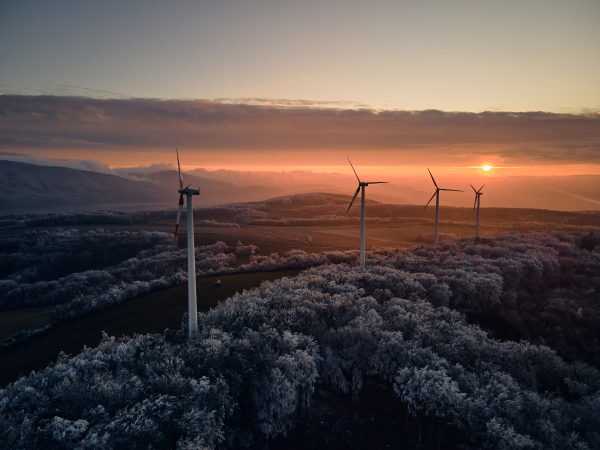 Aerial landscape photography of sunrise over frost-covered nature with wind turbines. Wind turbine towers in soft morning light with icy trees around, harmony of nature and technology. Concept of wind power as clean, renewable energy source.