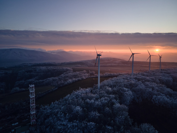 Aerial landscape photography of sunrise over frost-covered nature with wind turbines. Wind turbine towers in soft morning light with icy trees around, harmony of nature and technology. Concept of wind power as clean, renewable energy source.