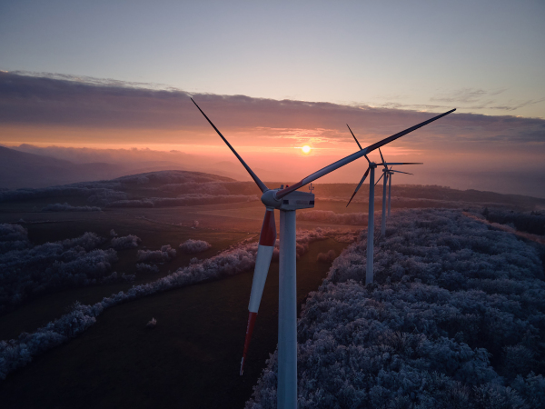 Aerial landscape photography of sunrise over frost-covered nature with wind turbines. Wind turbine towers in soft morning light with icy trees around, harmony of nature and technology. Concept of wind power as clean, renewable energy source.