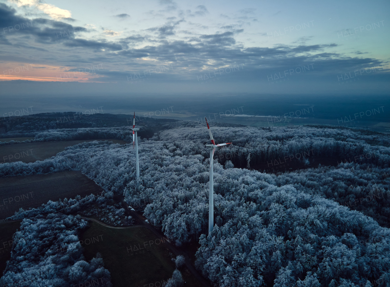 Aerial landscape photography of sunrise over frost-covered nature with wind turbines. Wind turbine towers in soft morning light with icy trees around, harmony of nature and technology. Concept of wind power as clean, renewable energy source.