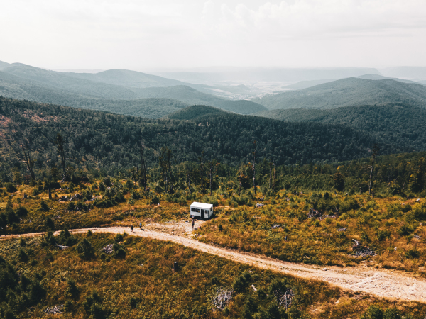 Aerial view of a small camper trailer in the middle of summer forest, with campers sitting by fire.