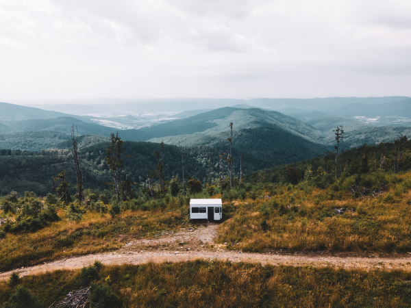 Aerial view of a small camper trailer in the middle of summer forest, with campers sitting by fire.