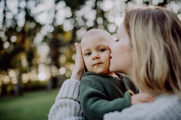 A mother holding and kissing her little baby son wearing knitted sweater during walk in nature, close-up