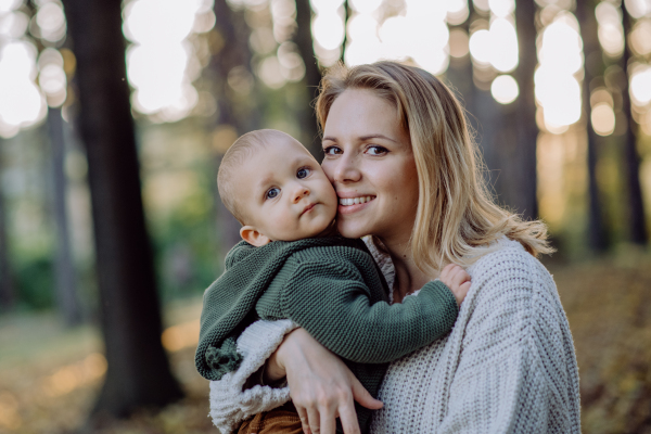 A mother holding her little baby son wearing knitted sweater during walk in nature, looking at camera.