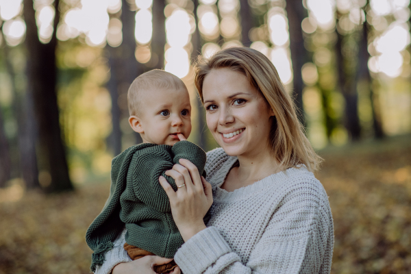 A mother holding her little baby son wearing knitted sweater during walk in nature, looking at camera.