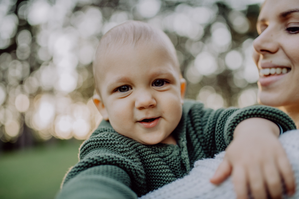 A mother holding her little baby son wearing knitted sweater during walk in nature, looking at camera.