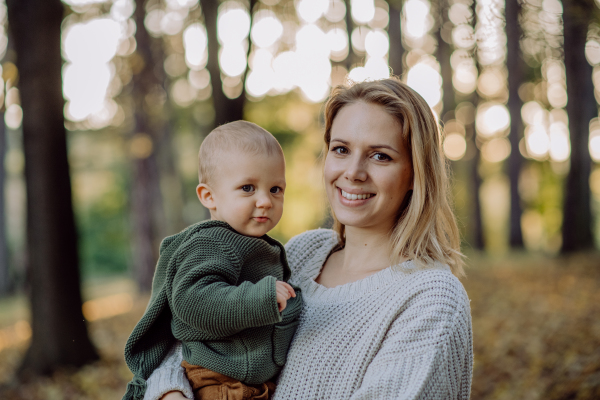 A mother holding her little baby son wearing knitted sweater during walk in nature, looking at camera.