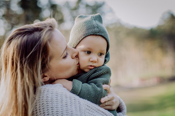 A mother holding and kissing her little baby son wearing knitted sweater during walk in nature, close-up