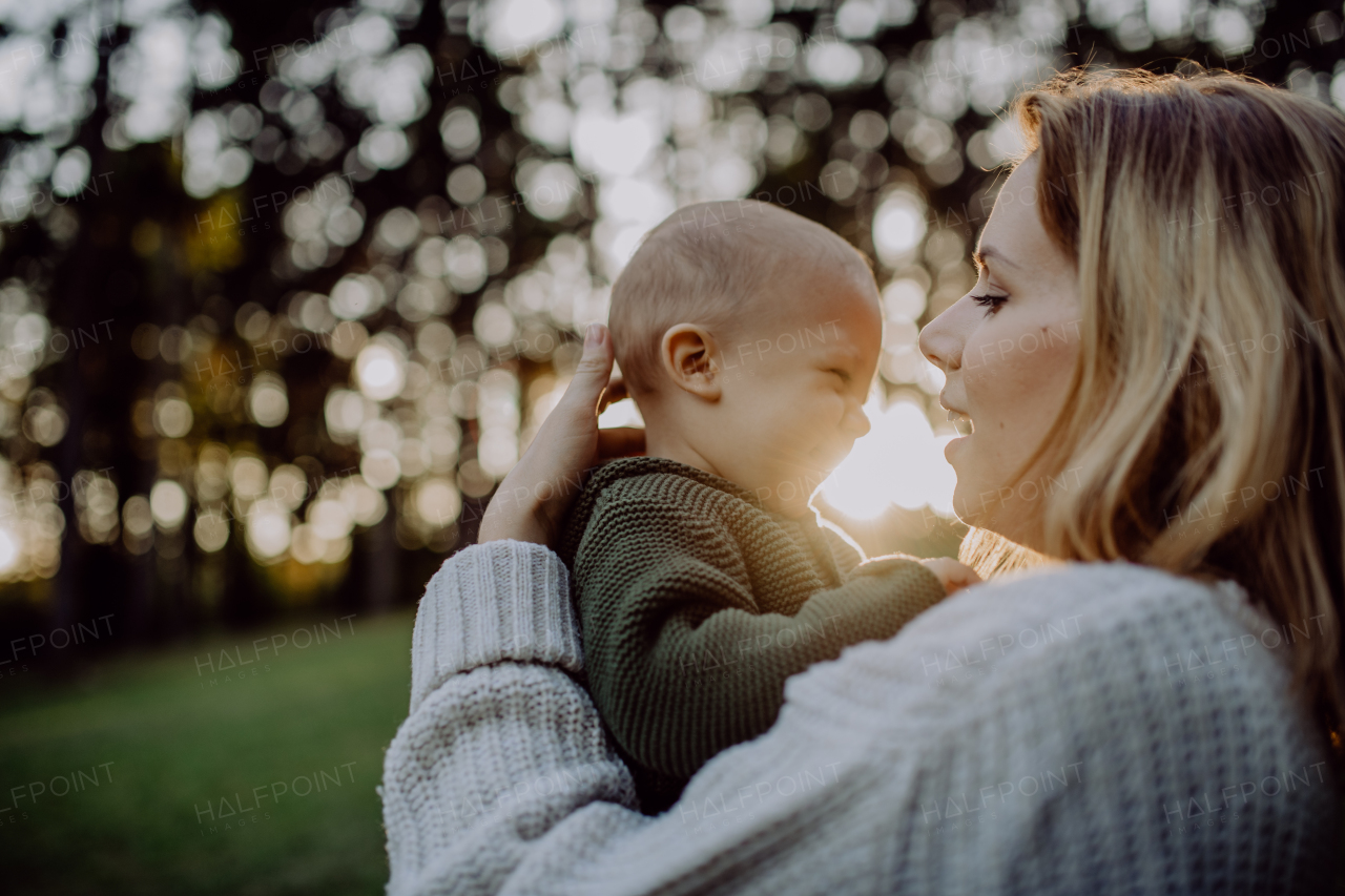 A mother holding her little baby son wearing knitted sweater during walk in nature, looking at camera.