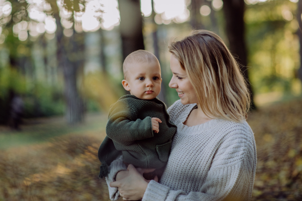 A mother holding her little baby son wearing knitted sweater during walk in nature, looking at camera.