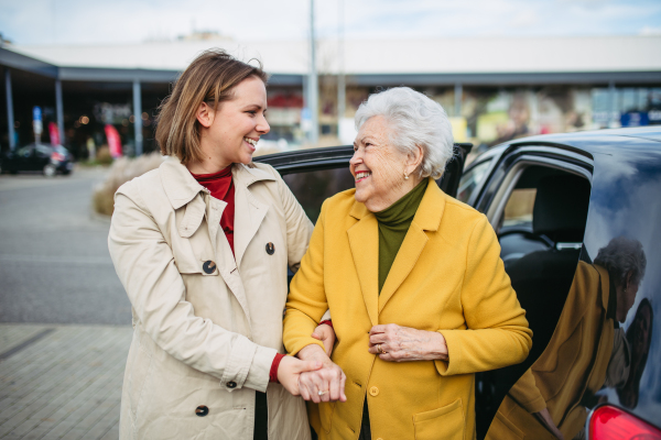 Senior lady getting out of the car, caregiver helping her, holding her hands. Elderly woman has problem with standing up from car back seat.