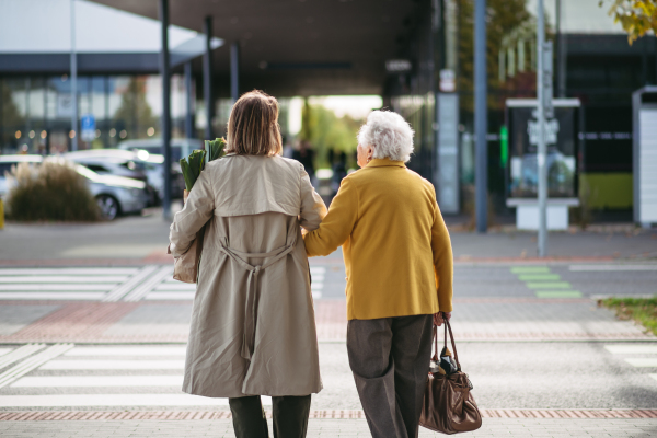 Rear view of mature granddaughter walking arm in arm from shop. Caregiver carrying groceries to the senior woman's car. Elderly lady shopping at the shopping center, needing help loading groceries into vehicle.