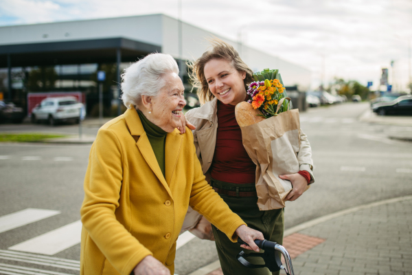 Mature granddaughter carrying grandmother's shopping bag. Senior woman and caregiver going to home with goceries from the supermarket, during cold autumn day.