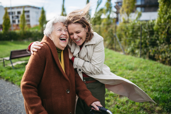 Grandmother and mature granddaughter on a walk in the city park, during a windy autumn day. Caregiver and senior lady enjoying the fall weather, laughing as the wind blows their hair.