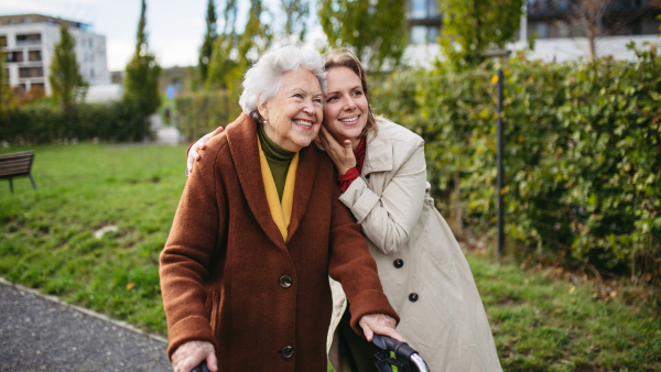 Front view of grandmother and mature granddaughter on a walk in the city park, during a windy autumn day. Caregiver and senior lady with mobility walker talking and enjoying the fall weather.