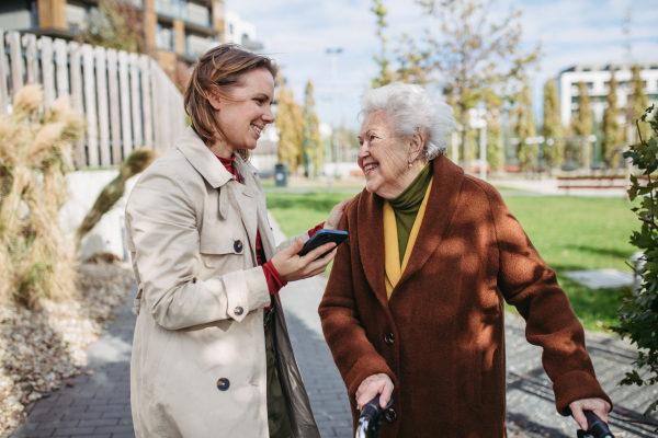 Front view of grandmother and mature granddaughter on a walk in the city park, during a windy autumn day. Caregiver and senior lady with mobility walker talking and enjoying the fall weather.