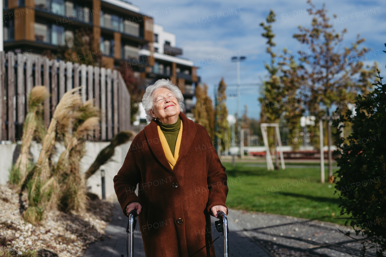 A senior woman with a mobility walker walking on the city streets during autumn day, enjoying the beautiful sunny weather. Elderly lady savoring every moment, living life to fullest.