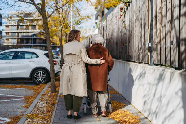 Rear view of grandmother and mature granddaughter on a walk in the city park, during a windy autumn day. Caregiver and senior lady with mobility walker enjoying the fall weather.